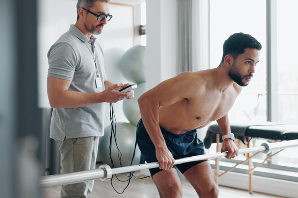 Man performing barbell exercise under clinical supervision while using REVO sports recovery device for physical therapy validation.