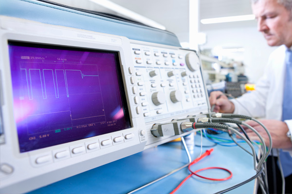Oscilloscope displaying voltage waveform on a test bench with an engineer working in the background.